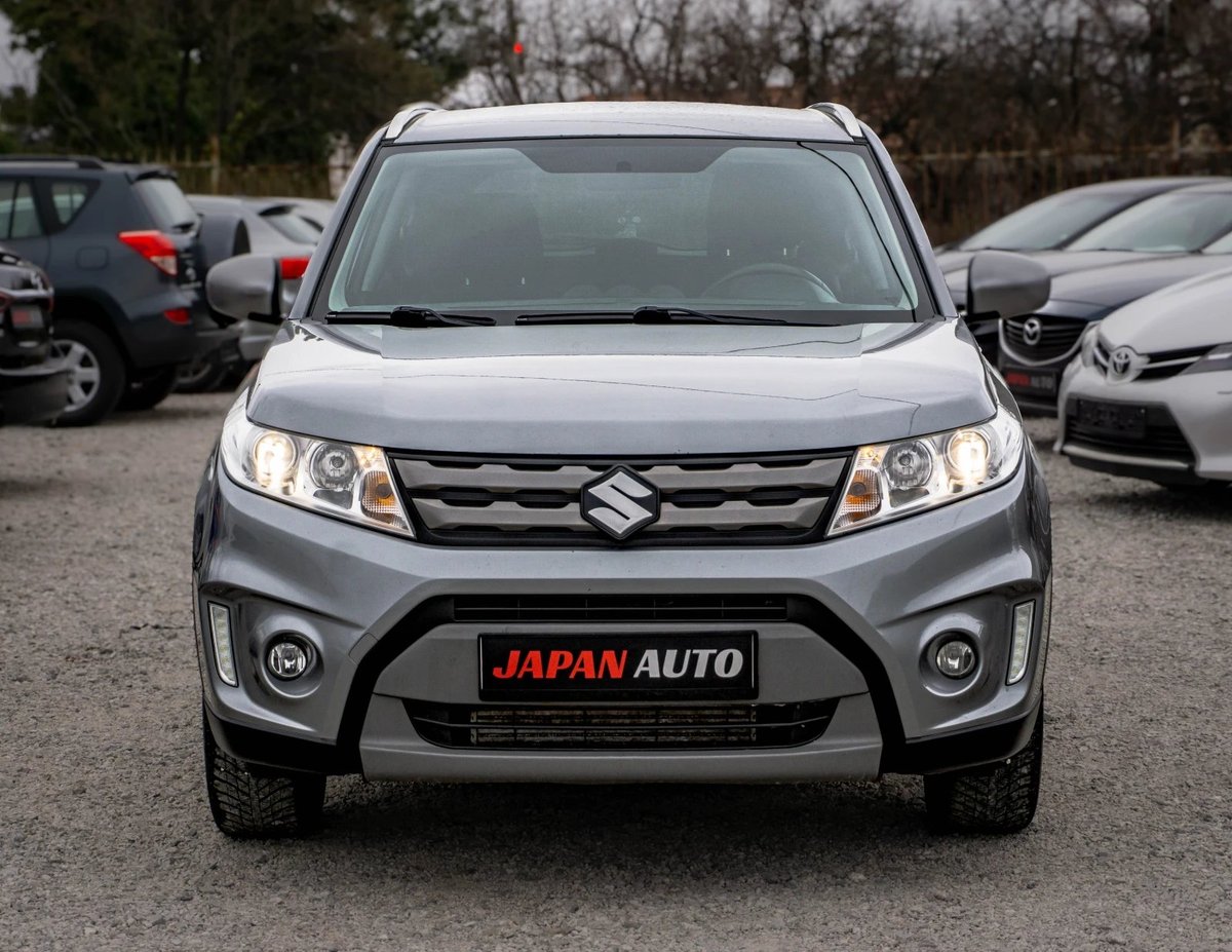 Silver Suzuki SUV parked in lot with Japan Auto dealership plate, front view showing grille and headlights