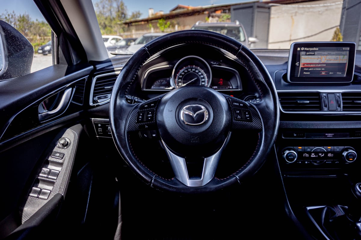 Interior view of Mazda vehicle dashboard featuring steering wheel, digital display, and controls with parking lot visible through windshield