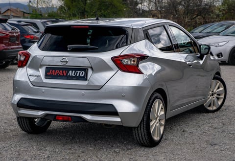 Silver Nissan hatchback car displayed at a dealership lot with other vehicles in the background