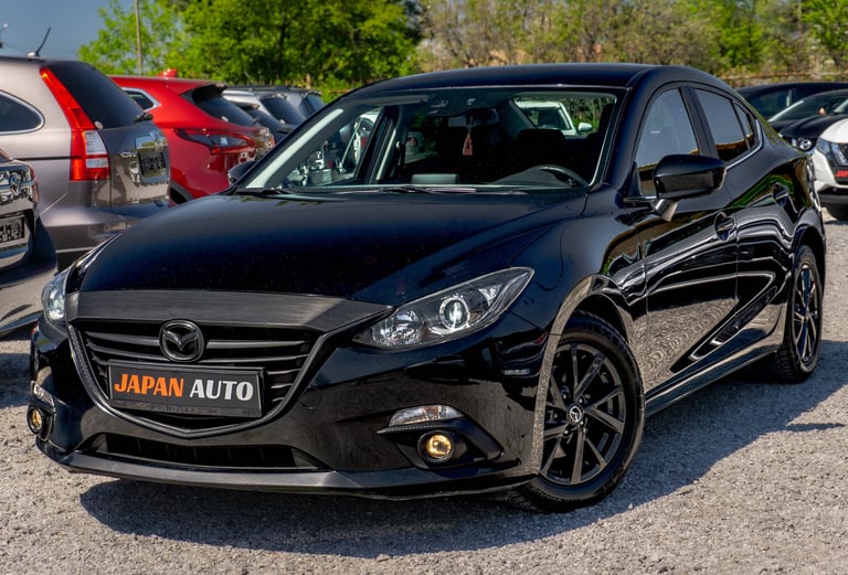 Black Mazda hatchback with Japan Auto badge parked in a lot with other vehicles and trees in background
