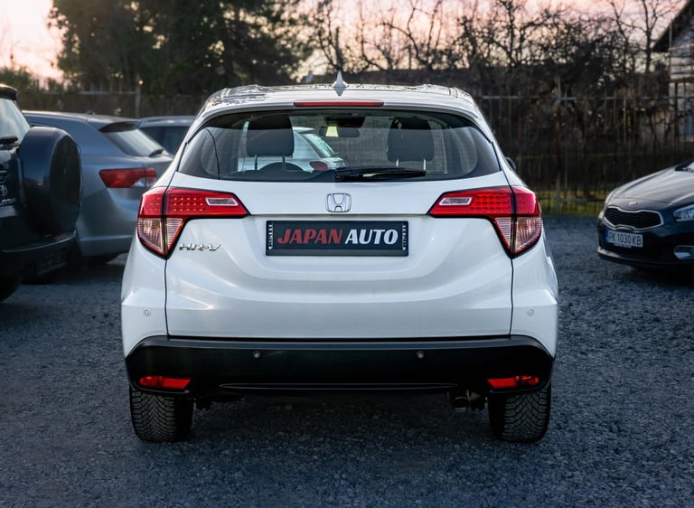 White hatchback car viewed from rear in a dealership lot with bare trees in background