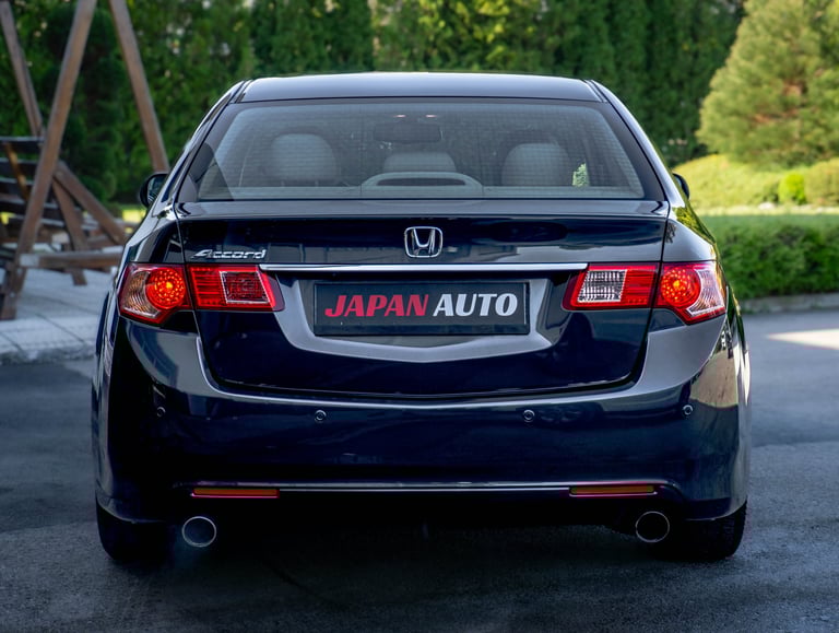 Black Honda sedan viewed from rear, showing illuminated red taillights and Japan Auto dealership plate, parked outdoors with trees in background