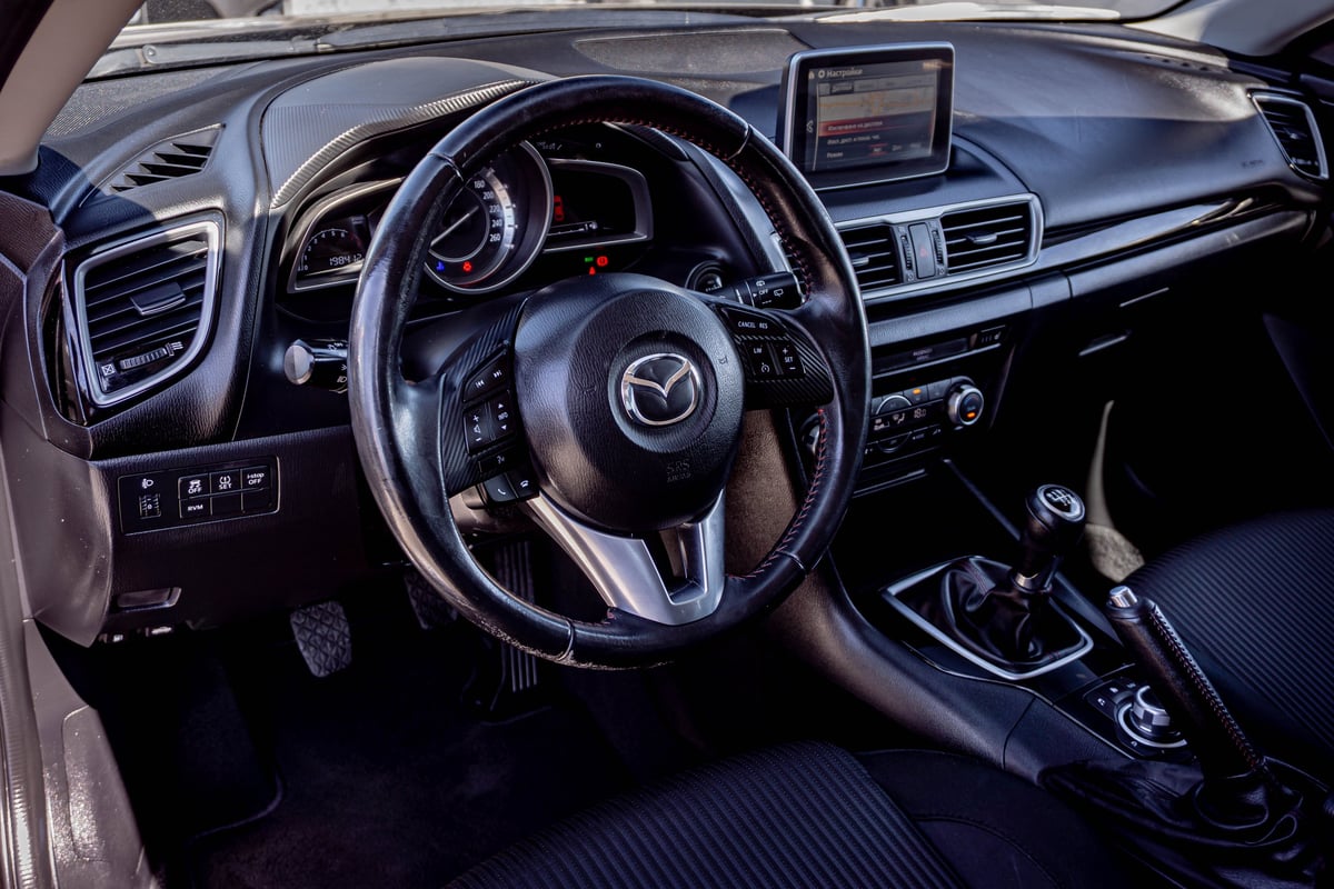 Modern car interior dashboard featuring a black leather steering wheel with Mazda logo, digital instrument cluster, touchscreen display, and center console with manual transmission