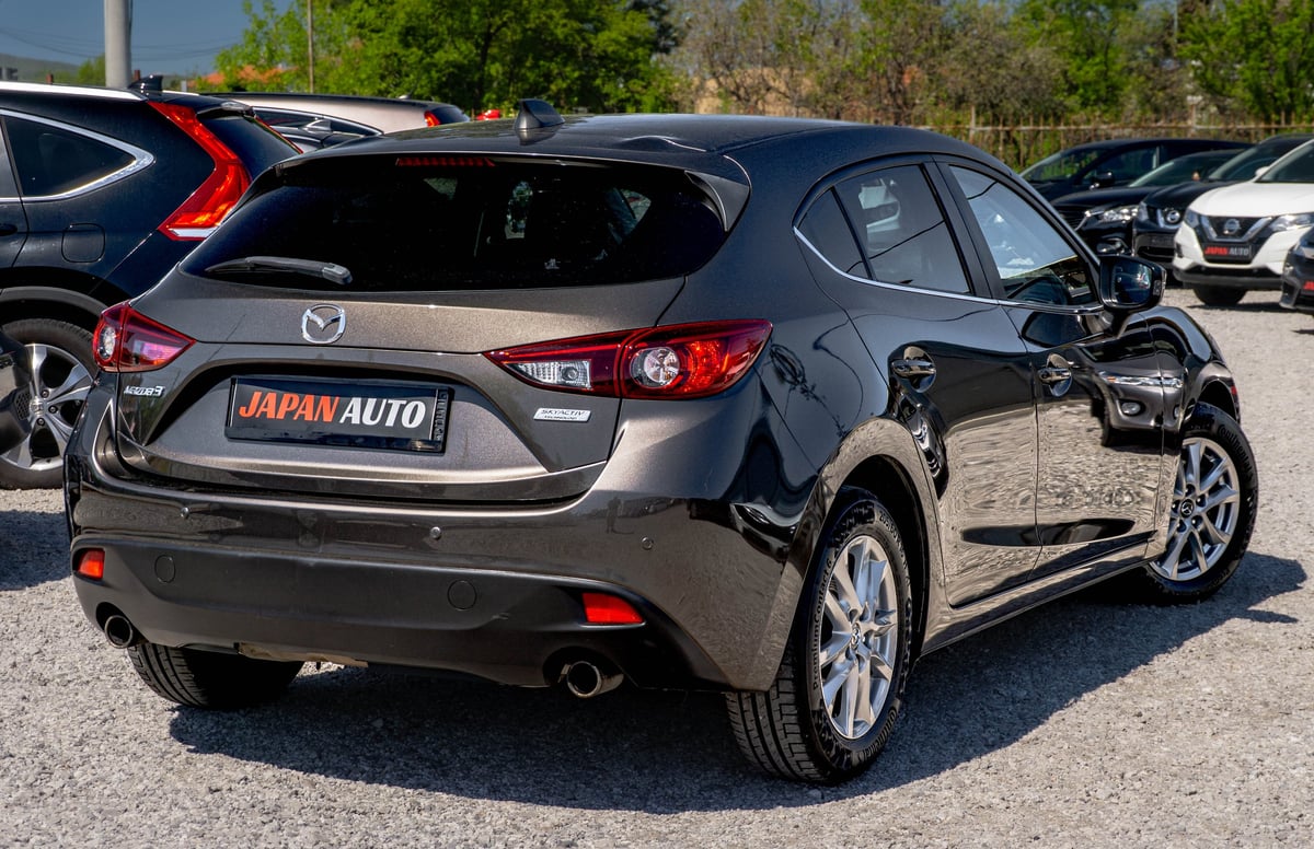 Gray Mazda hatchback car with Japan Auto dealer plate parked in outdoor lot with other vehicles visible