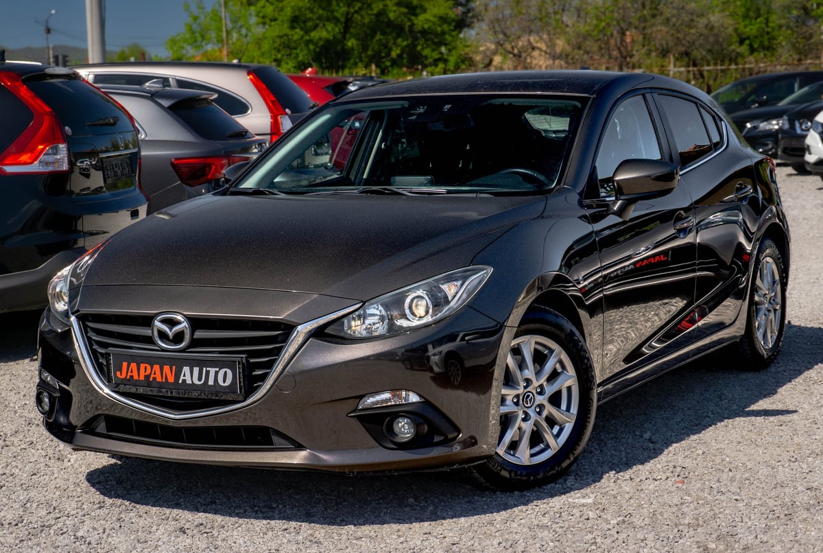 Black Mazda compact car with Japan Auto dealership plate displayed in parking lot with other vehicles visible in background