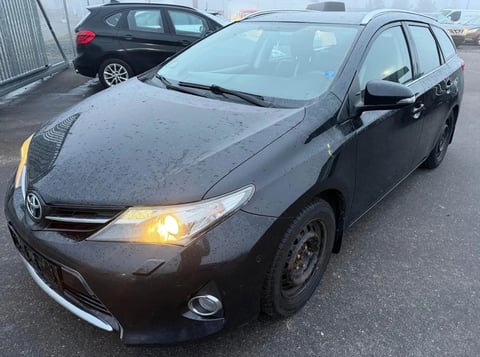 Black Toyota Auris hatchback parked in a lot with headlights on, showing wet surface and another car in the background