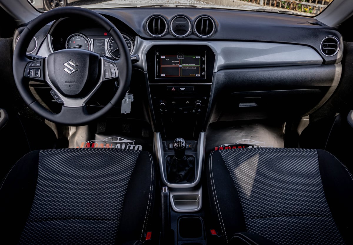 Interior of a Suzuki vehicle with black steering wheel, dashboard, touchscreen display, and black and gray upholstered seats