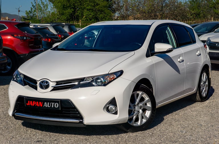 White Toyota hatchback car with JAPAN AUTO license plate parked in a lot with other vehicles and trees in background