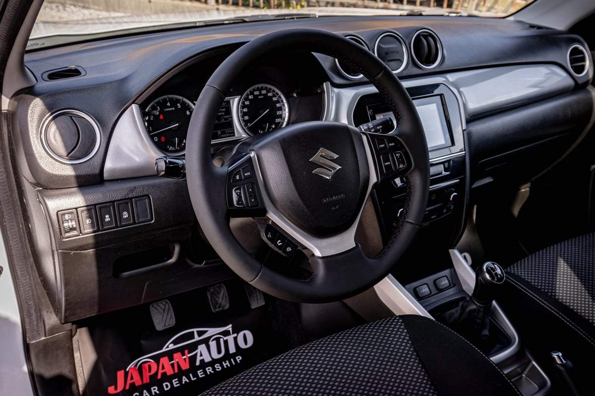 Interior dashboard and steering wheel of a Suzuki vehicle, showing black leather steering wheel, instrument cluster, and control panel