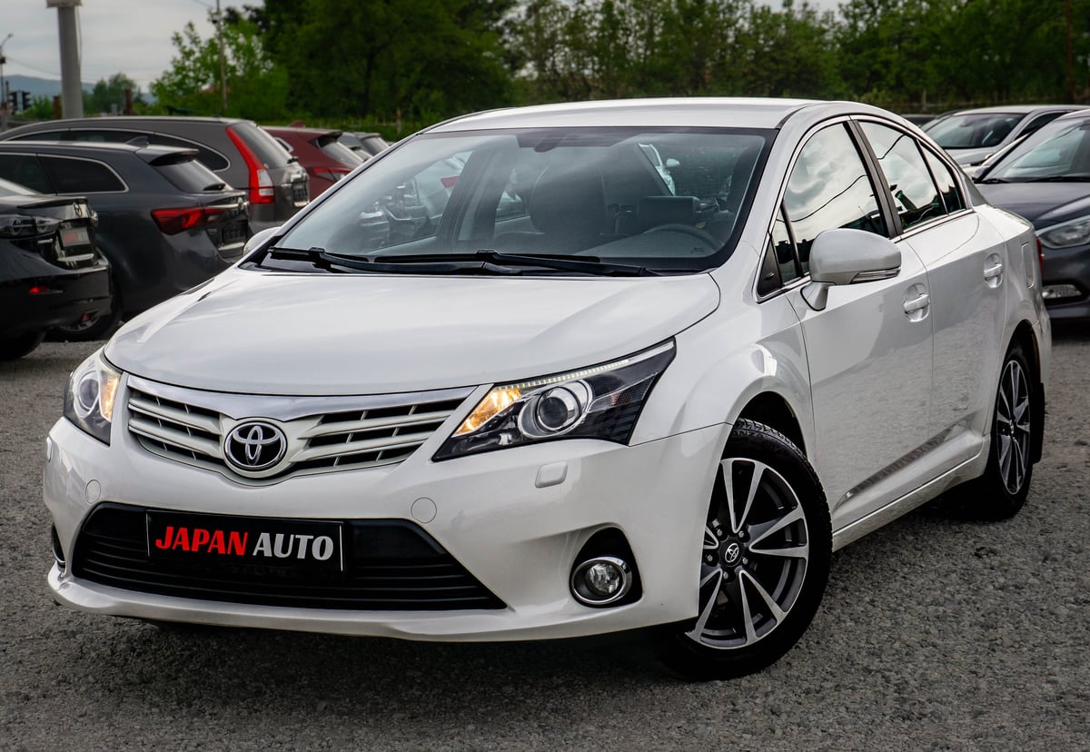White Toyota Avensis sedan parked in a lot with other vehicles, featuring alloy wheels and Japan Auto branding