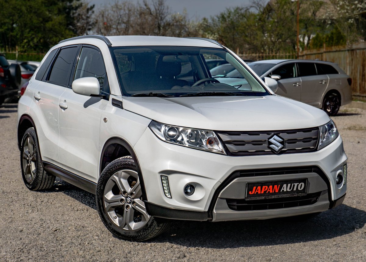 White Suzuki SUV with Japan Auto badge parked at a dealership lot with trees and vehicles in the background